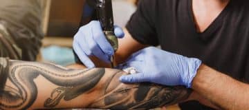 close-up-professional-tattooer-artist-doing-tattoo-arm-young-man-by-machine-with-black-ink-1200x520